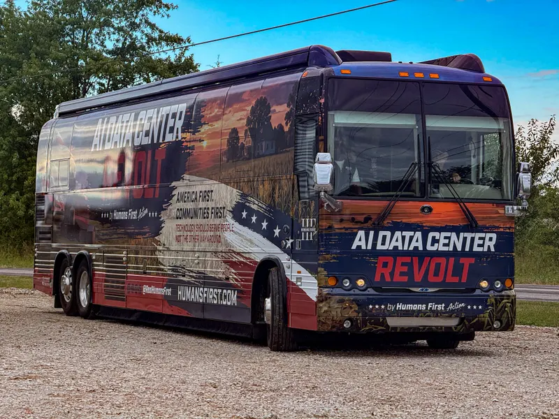 A tour bus wrapped in red, white and blue graphics reading "AI Data Center Revolt by Humans First Action" is parked on a gravel lot. The side shows a farmhouse and cornfield at sunset with the slogans "America First. Communities First." and "Technology should serve people, not the other way around," along with HUMANSFIRST.COM. Trees and a blue sky are in the background.