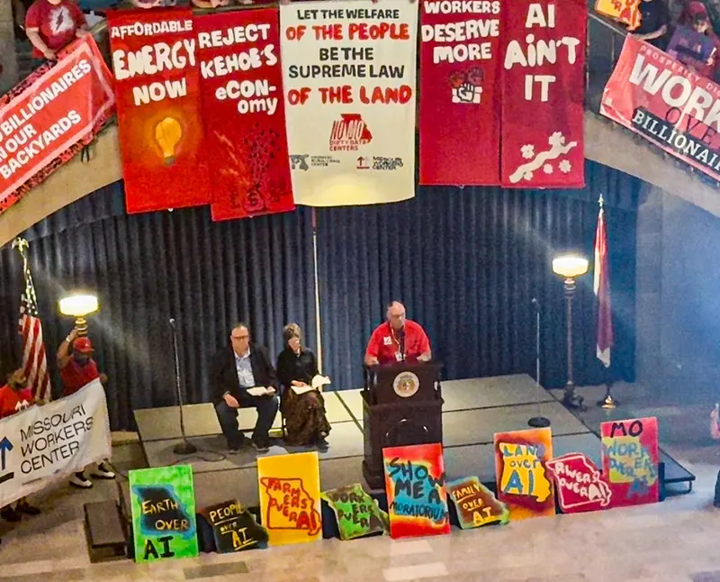 Seen from above, a farmer in a red shirt speaks at a podium bearing the Missouri state seal on a stage in the Capitol rotunda, while a man and a woman sit behind him holding papers. Red and white banners hang overhead, reading "Affordable Energy Now," "Reject Kehoe's Econ-omy," "Let the Welfare of the People Be the Supreme Law of the Land," "Workers Deserve More" and "AI Ain't It." Hand-painted signs shaped like the state of Missouri line the front of the stage, reading "Earth Over AI," "People Over AI," "Farmers Over AI," "Show Me A Moratorium," "Land Over AI" and "Rivers Over AI." A Missouri Workers Center banner is held at left beside an American flag.
