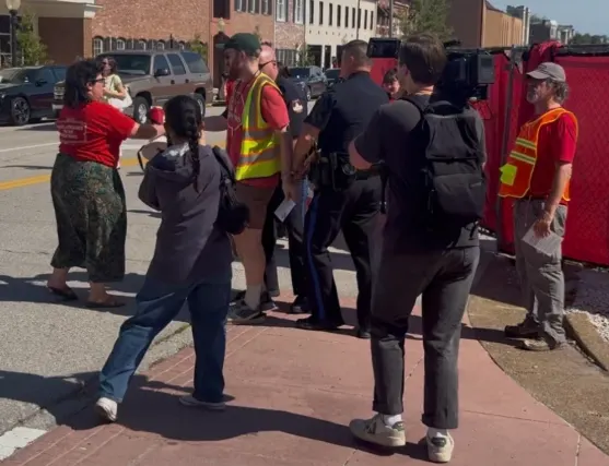 A Jefferson City police officer escorts a man in a green cap, red T-shirt and yellow safety vest along a brick sidewalk during a data center protest. A woman in a red shirt reaches toward them from the street, and a young woman in a dark hoodie steps forward. A camera operator films the arrest while a man in an orange safety vest watches beside a red construction fence.