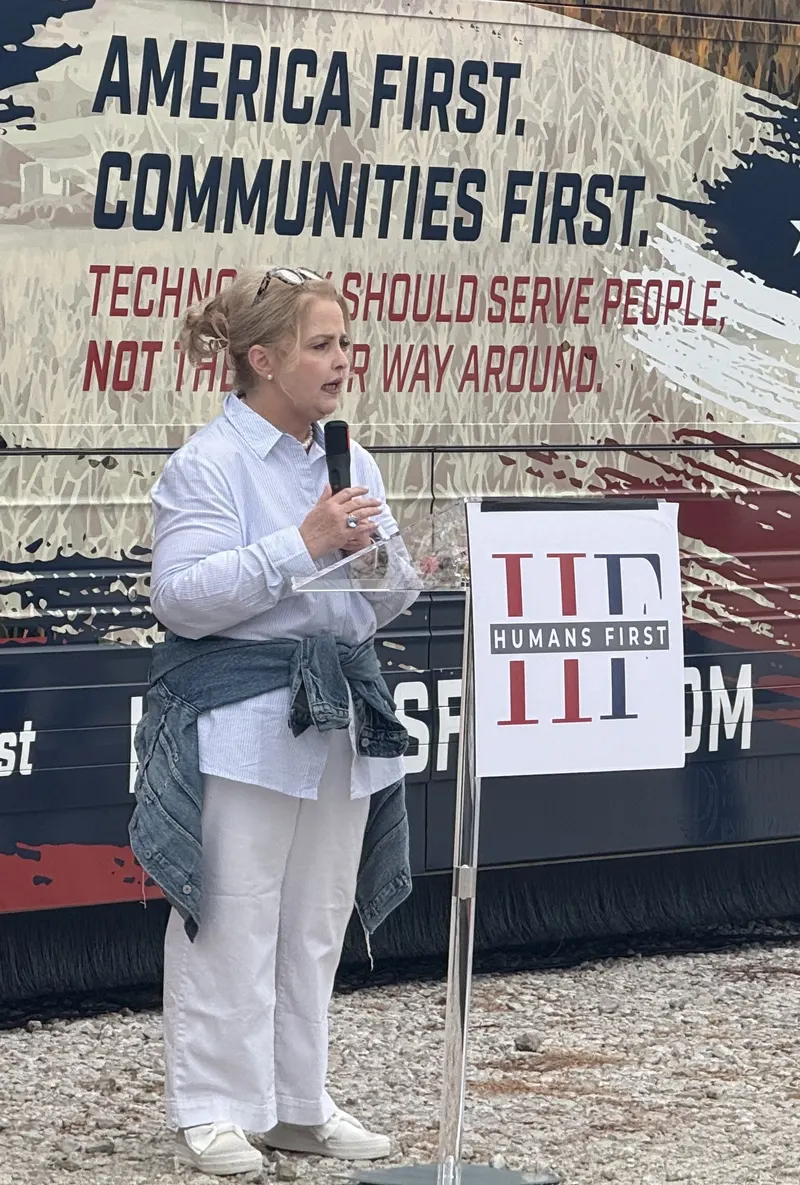Amy Kremer, in a light blue striped shirt and white pants with a denim jacket tied at her waist, speaks into a handheld microphone beside a clear podium with a Humans First sign. Behind her, the tour bus wrap reads "America First. Communities First. Technology should serve people, not the other way around."