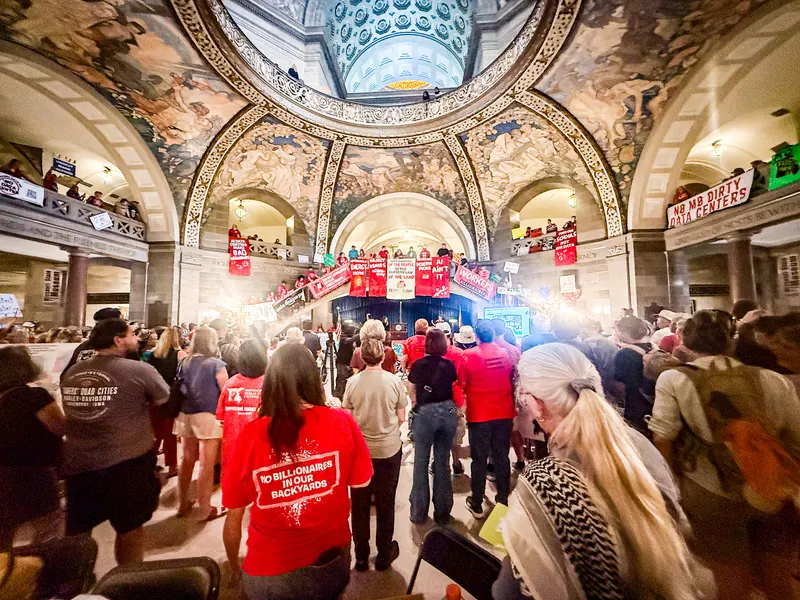 A wide-angle view from inside the crowd in the Missouri Capitol rotunda, under the painted dome and murals. A woman in the foreground wears a red T-shirt reading "No Billionaires in Our Backyards." Protesters in red shirts line the grand staircase and balconies beneath red banners, including "AI Ain't It," "Workers Deserve More," "Reject Kehoe's Con-omy" and "No Mo Dirty Data Centers." A speaker stands at a podium at the foot of the stairs.