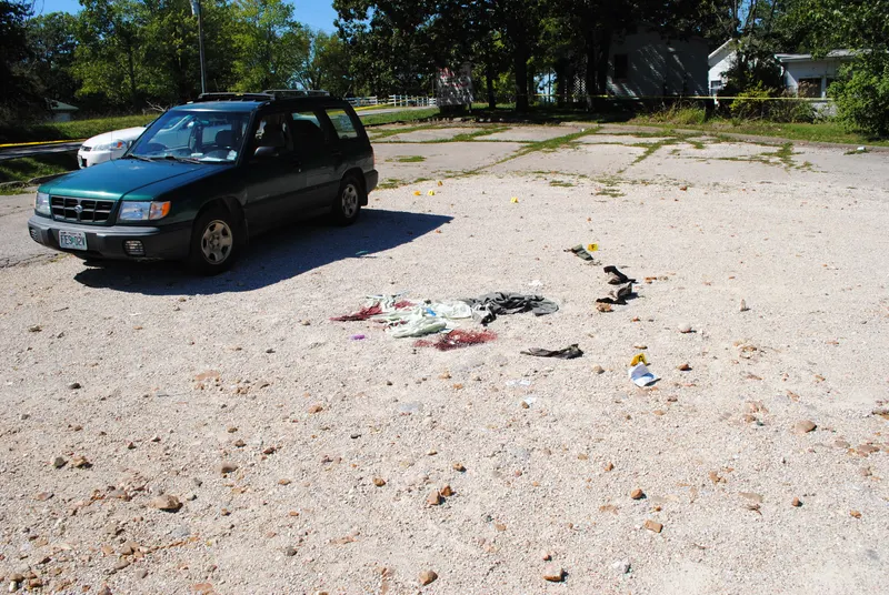 Missouri State Highway Patrol crime scene photograph of the September 11, 2012 shooting scene, showing Jeff Weinhaus’s green Subaru Forester, scattered evidence, and his pistol partially withdrawn from an unsecured holster, with the barrel pointing south, opposite its apparent northward orientation in the watch camera footage. Missouri State Highway Patrol crime scene photograph of the September 11, 2012 shooting scene, showing Jeff Weinhaus’s green Subaru Forester, scattered evidence, and his pistol partially withdrawn from an unsecured holster, with the barrel pointing south, opposite its apparent northward orientation in the watch camera footage.