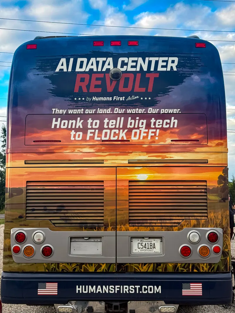 The back of the AI Data Center Revolt tour bus, wrapped in a farm-field sunset scene. The text reads "AI Data Center Revolt by Humans First Action," "They want our land. Our water. Our power." and "Honk to tell big tech to FLOCK OFF!" Across the bumper, HUMANSFIRST.COM appears between two American flags.