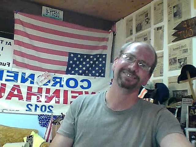 Jeff Weinhaus smiling during one of his 2012 broadcasts, with an American flag and his campaign banner behind him